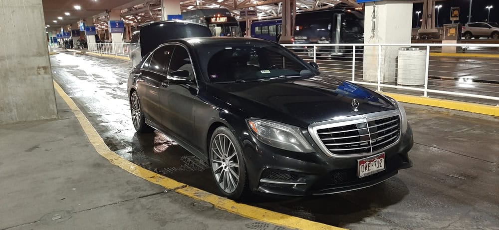 A black Mercedes-Benz sedan is parked under a covered area at night, reflecting the wet pavement beneath it.